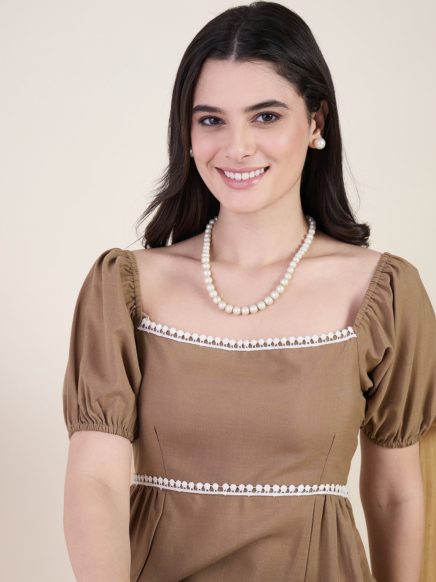 Woman wearing a brown dress with white trim and a pearl necklace against a beige background