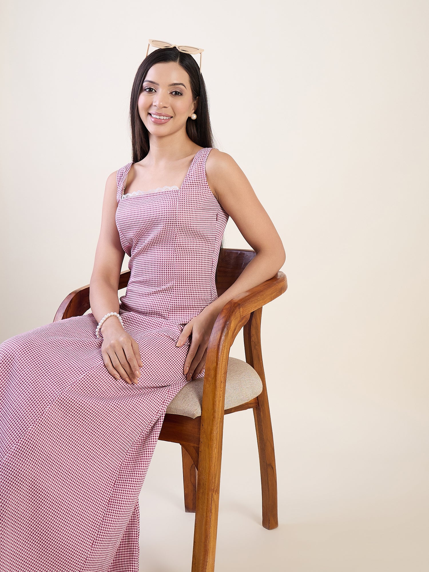 Woman wearing a pink checkered dress sitting on a wooden chair against a plain background
