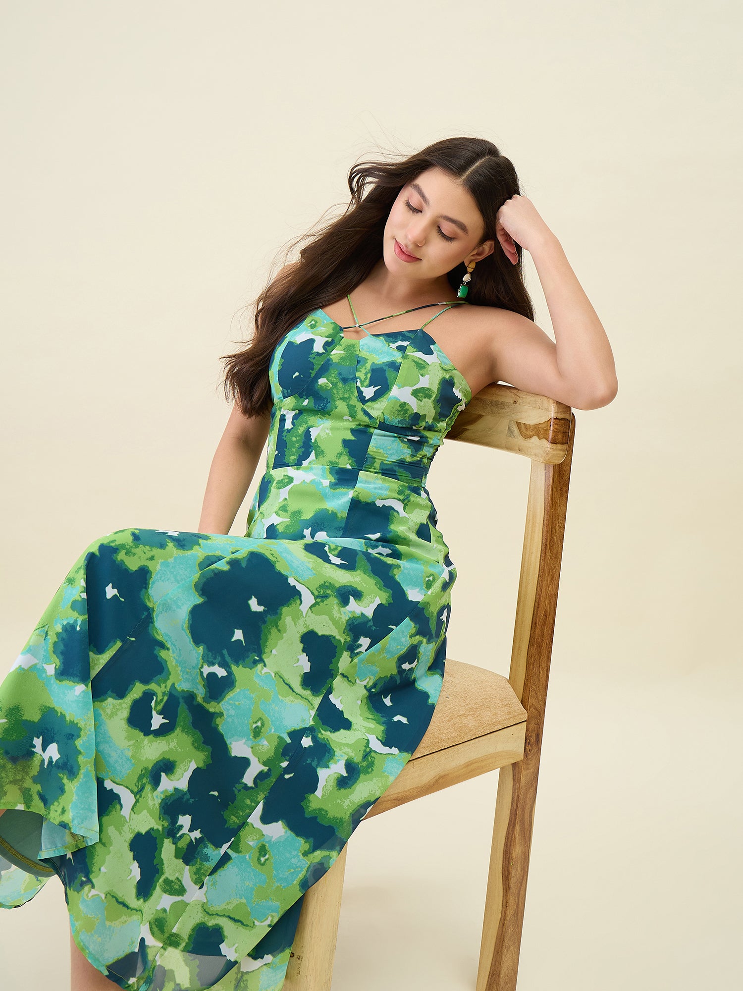 Woman wearing a green floral dress sitting on a wooden chair against a beige background