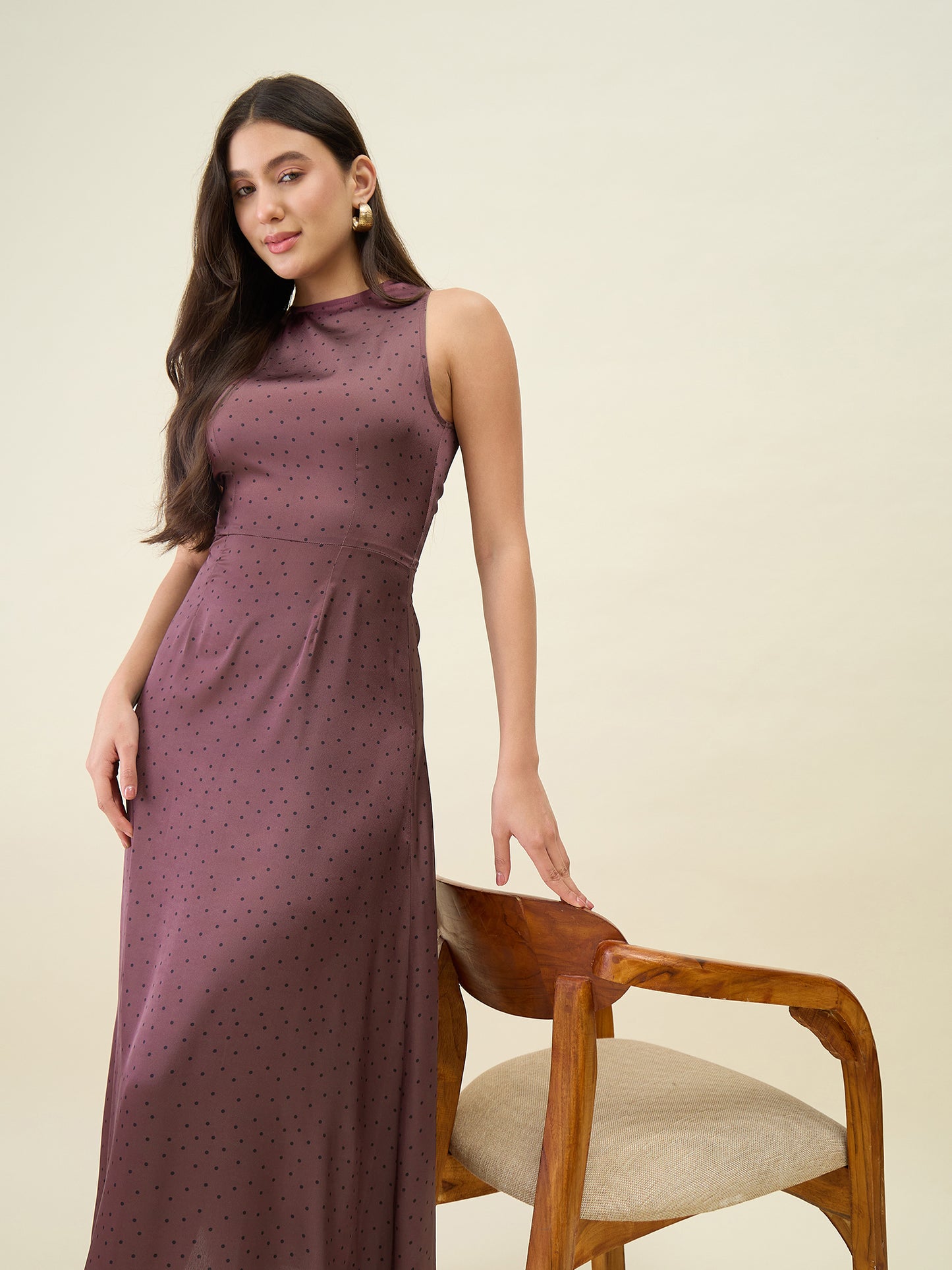 Woman wearing a long brown dress sitting on a wooden chair against a beige background