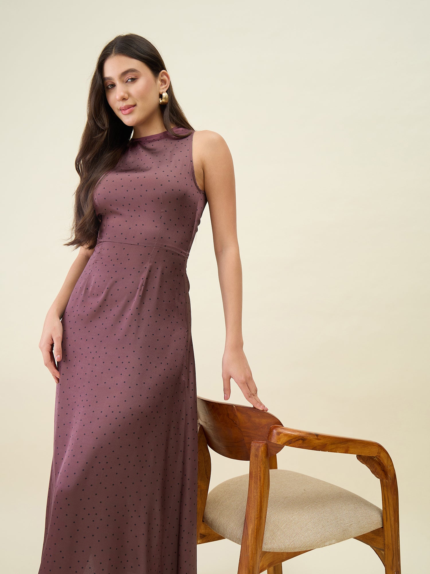 Woman wearing a long brown dress sitting on a wooden chair against a beige background