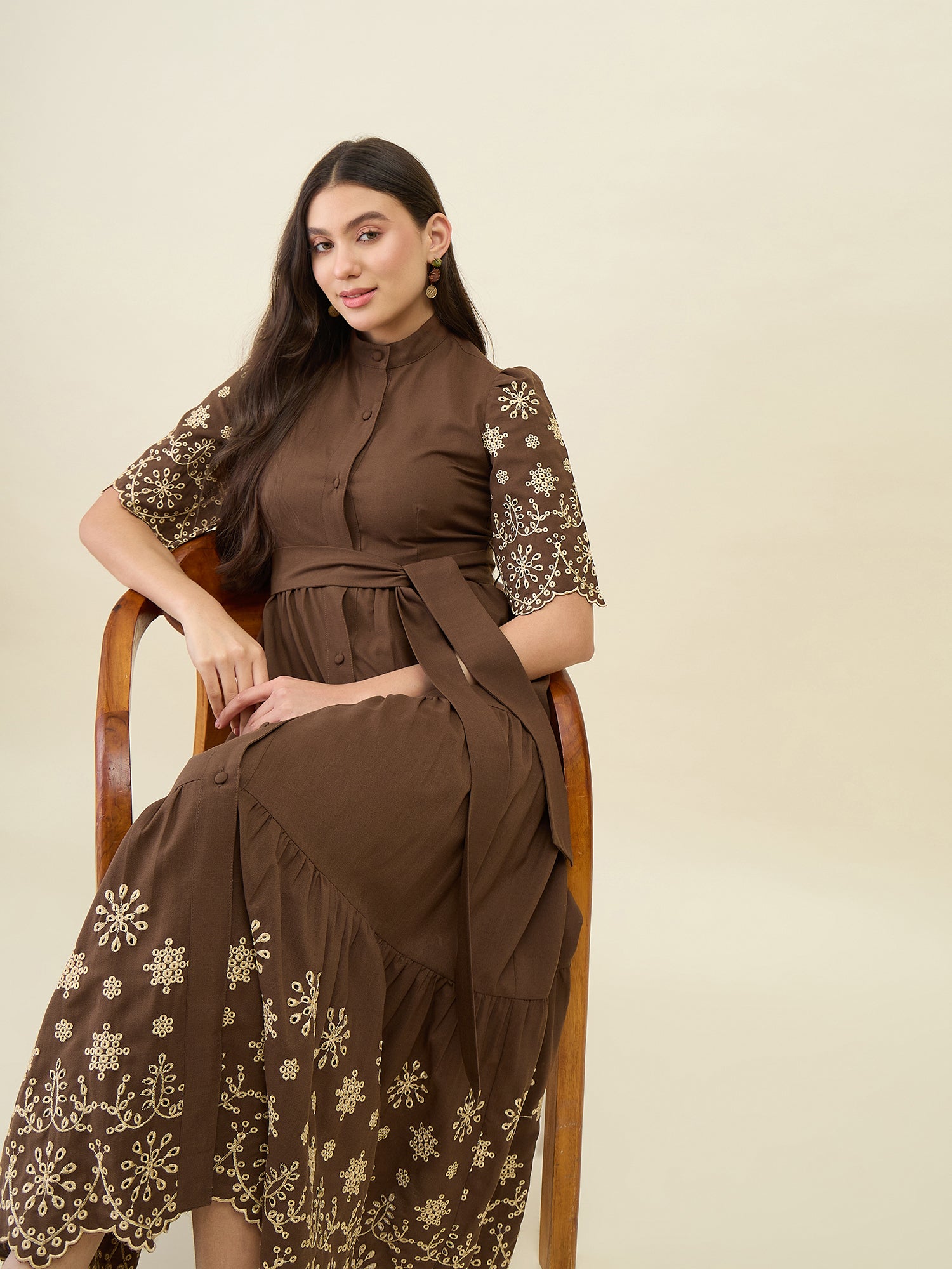 Woman wearing a brown dress with floral embroidery sitting on a chair against a beige background