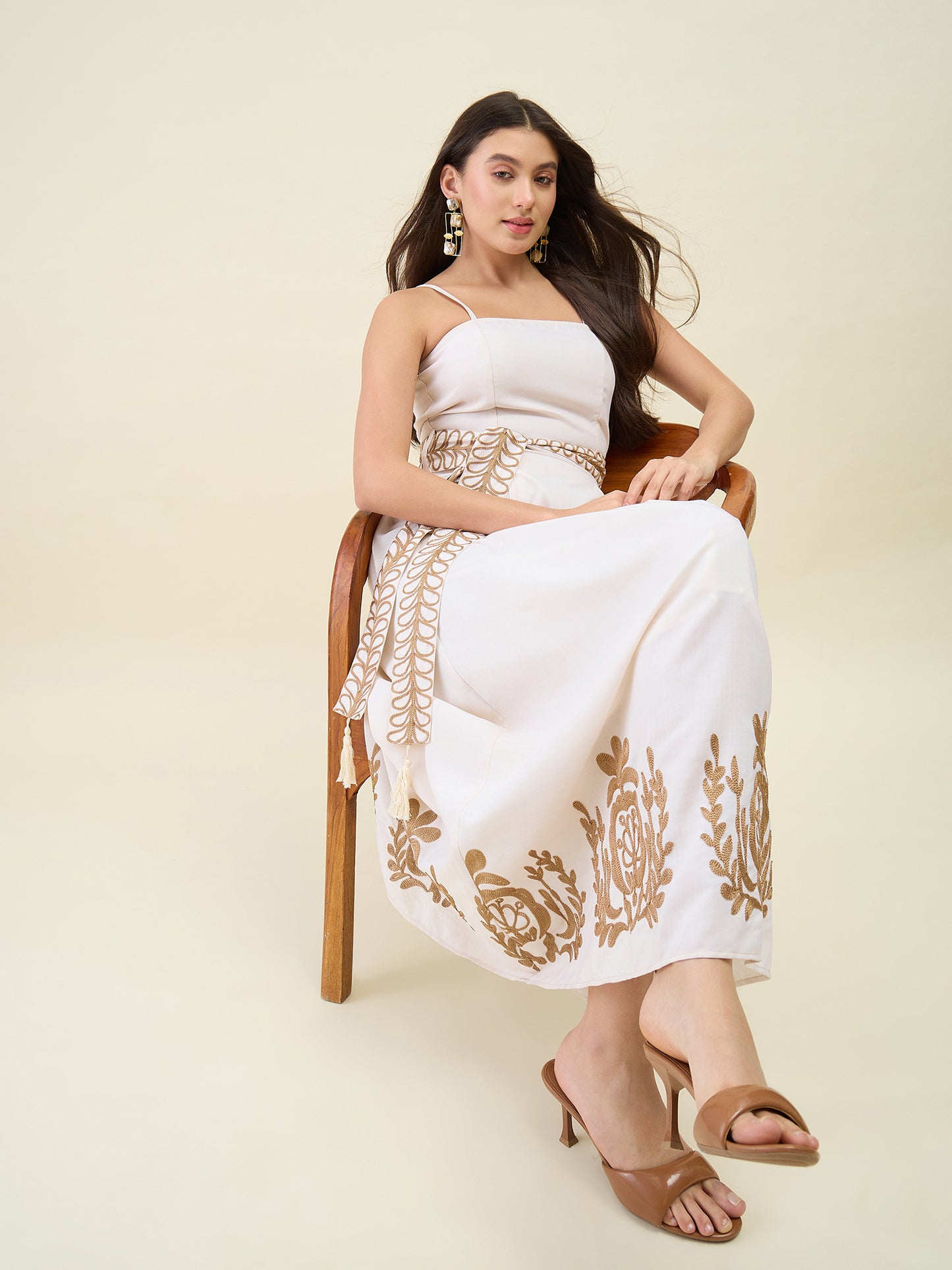 Woman wearing a white saree with gold embroidery sitting on a chair against a beige background