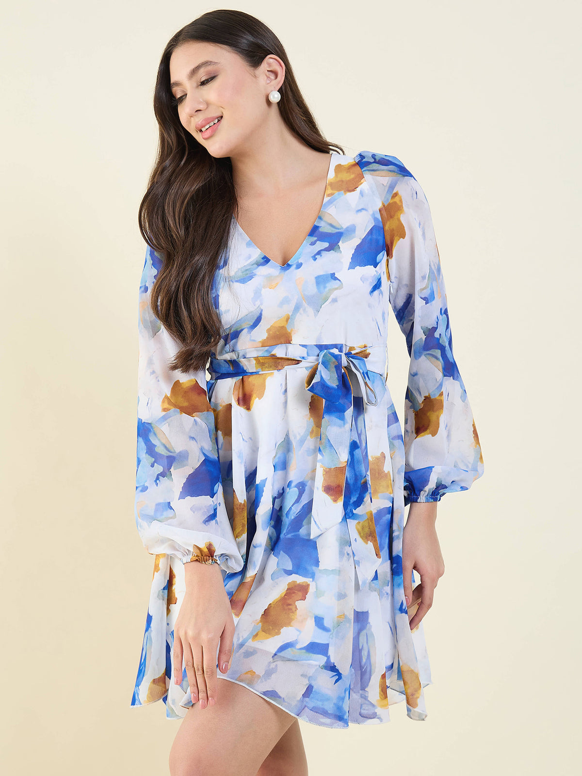 A woman modeling a short V-neck dress with a blue, white, and brown abstract watercolor floral print, featuring balloon sleeves and a matching tie belt at the waist, set against a solid light beige background.