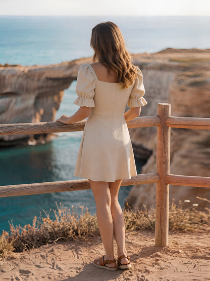 Woman in a beige dress standing on a cliff overlooking the ocean.