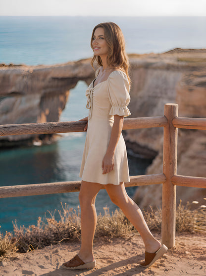 Woman in a beige dress standing by a wooden railing with a scenic background.