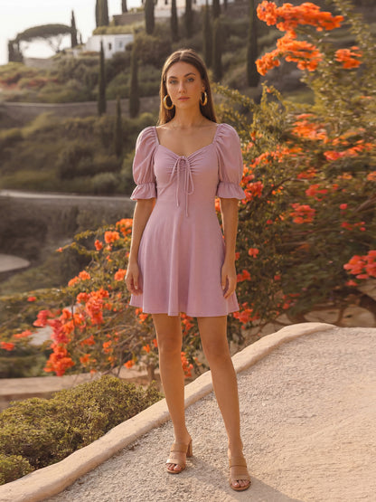 Woman in a mauve dress standing in a scenic outdoor setting with flowers and greenery.