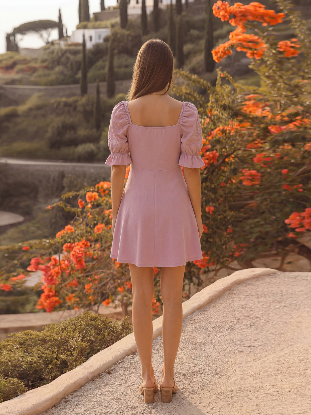 Woman in a mauve dress standing on a path with orange flowers and greenery around.