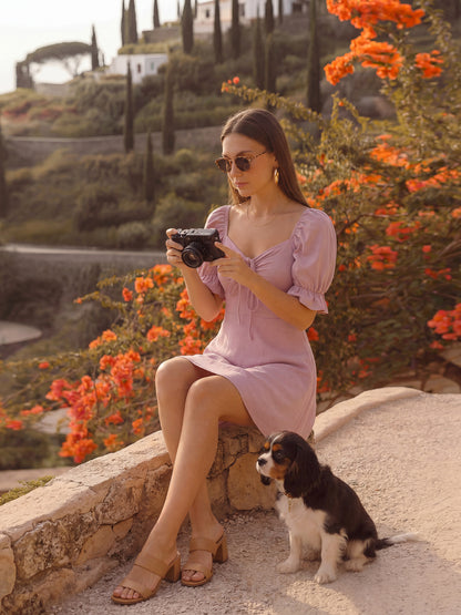 Woman in a mauve dress taking a photo with a camera, sitting on a stone ledge with a dog next to her, surrounded by orange flowers and greenery.