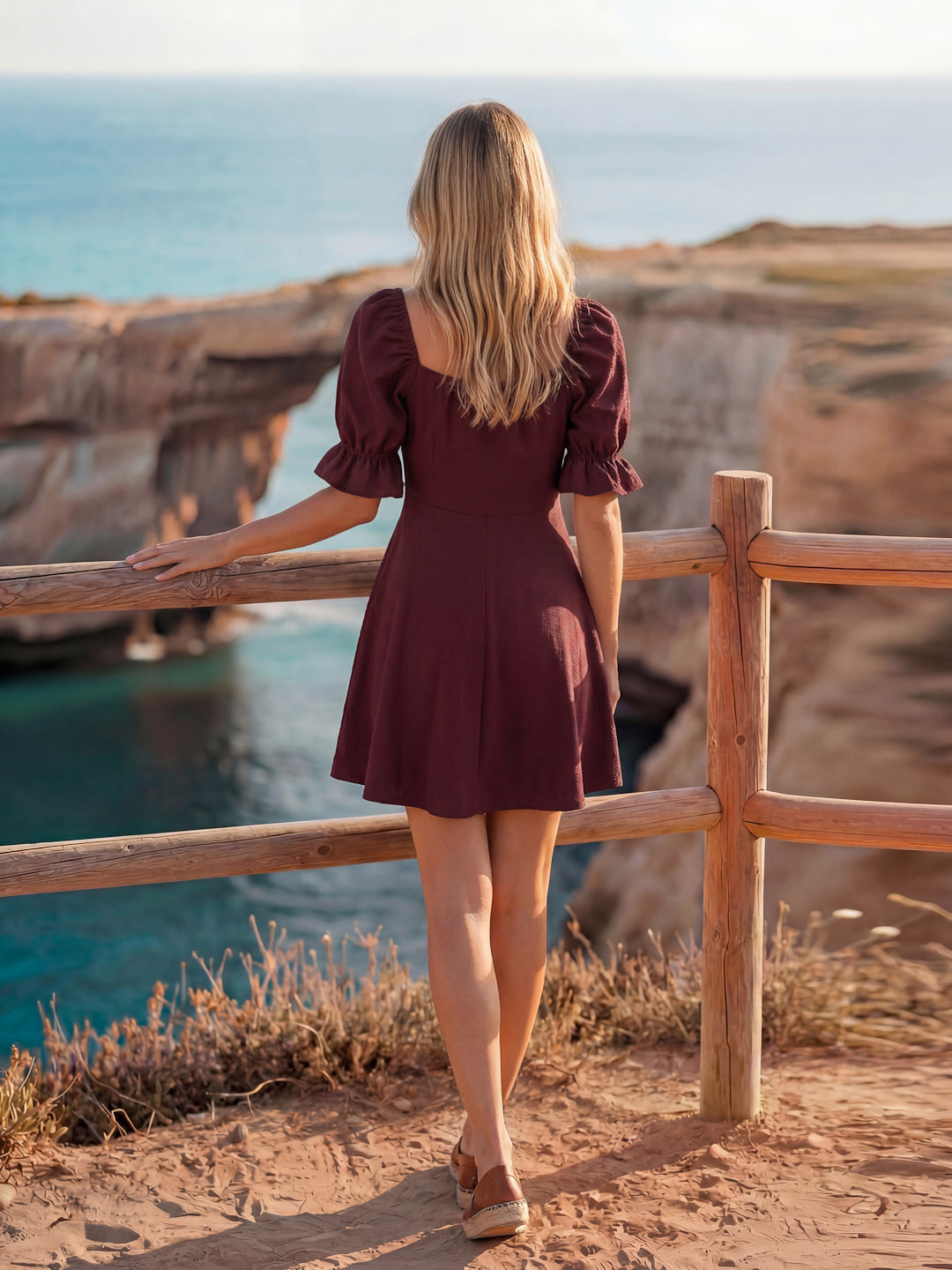 Woman in a burgundy dress standing on a cliff overlooking the ocean.