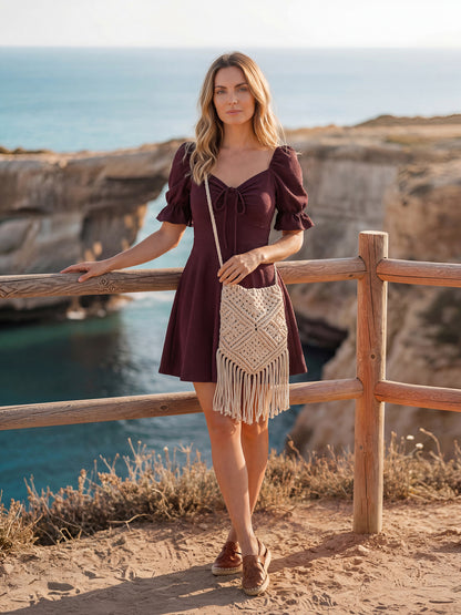 Woman in a wine color dress with a beige bag standing on a cliff overlooking the ocean.