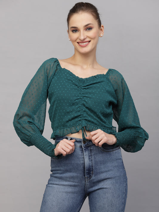 Woman wearing a green top with sheer sleeves and blue jeans on a gray background