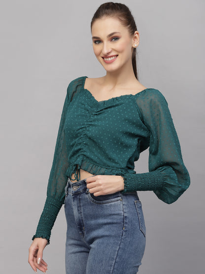 Woman wearing a green top with sheer sleeves and blue jeans on a gray background
