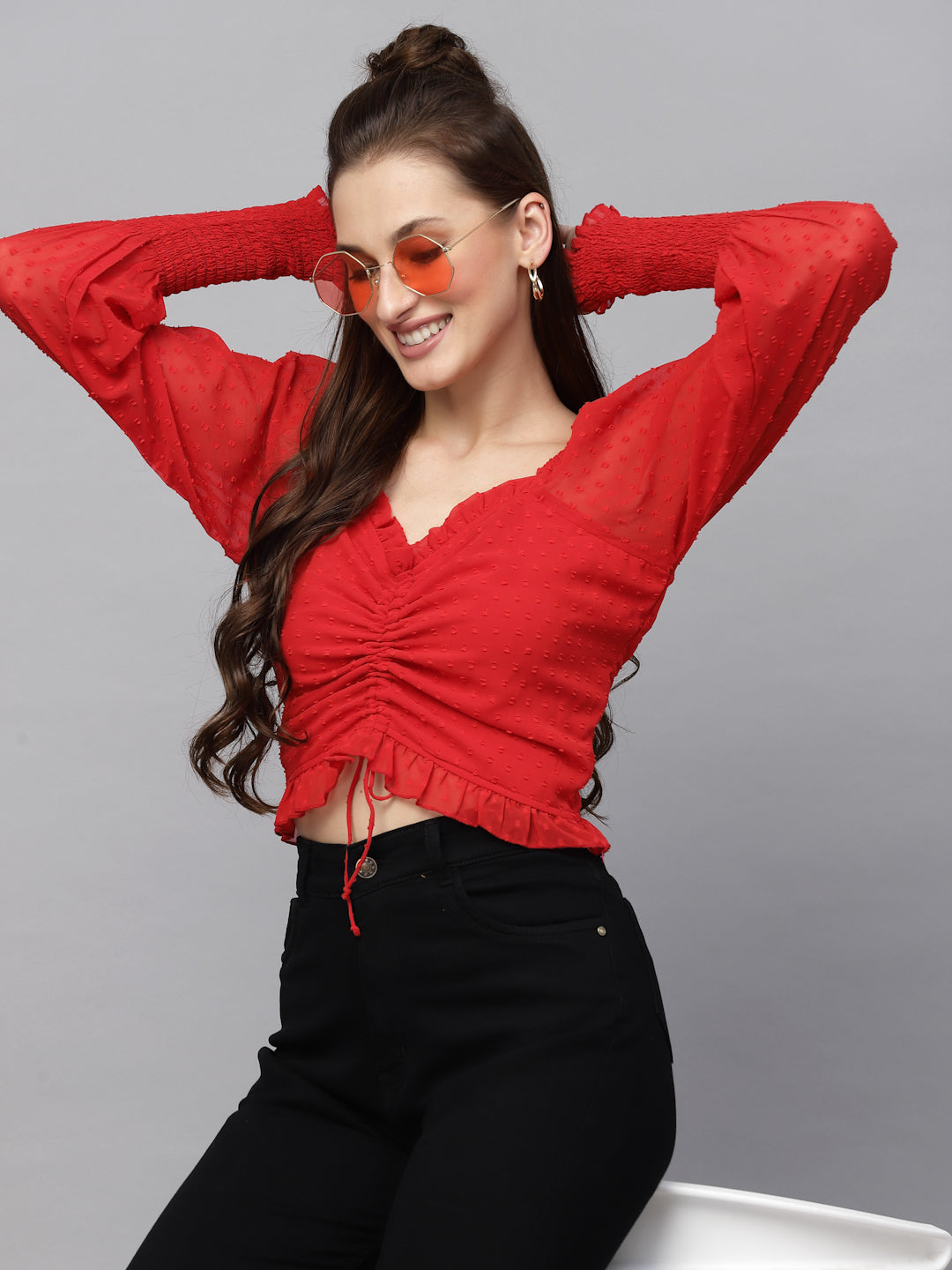 Woman wearing a red top and black pants with orange sunglasses against a gray background