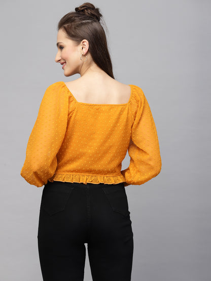 Woman wearing a mustard yellow top with long sleeves and black pants on a gray background