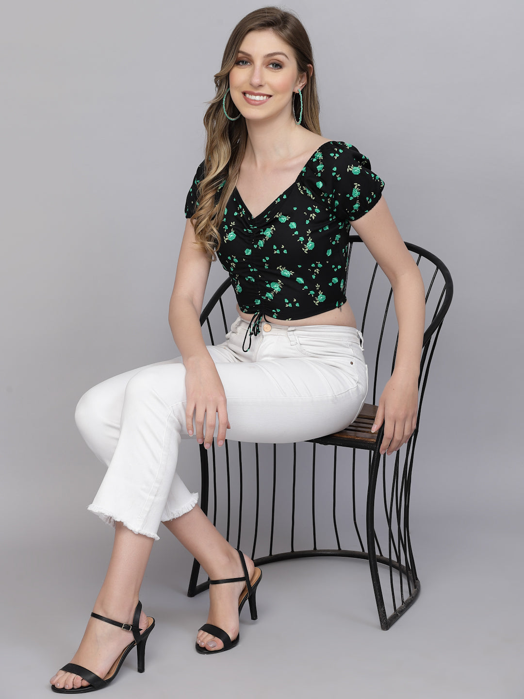 Woman sitting on a black chair wearing a black top with green patterns and white pants.