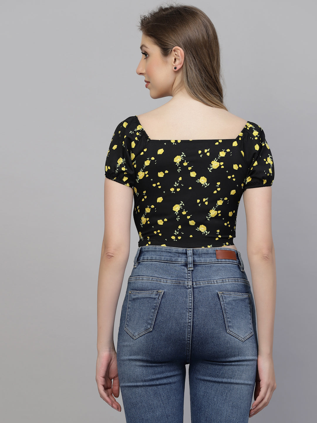Woman wearing a black top with yellow floral patterns and blue jeans on a gray background