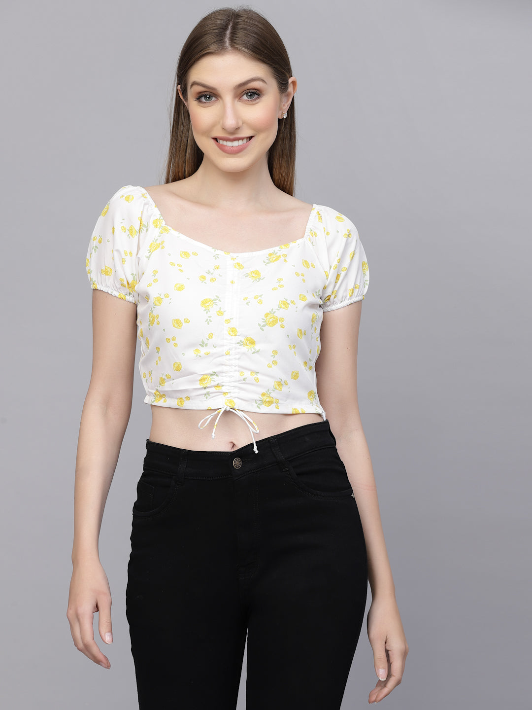 Woman wearing a white crop top with yellow patterns and black pants on a gray background