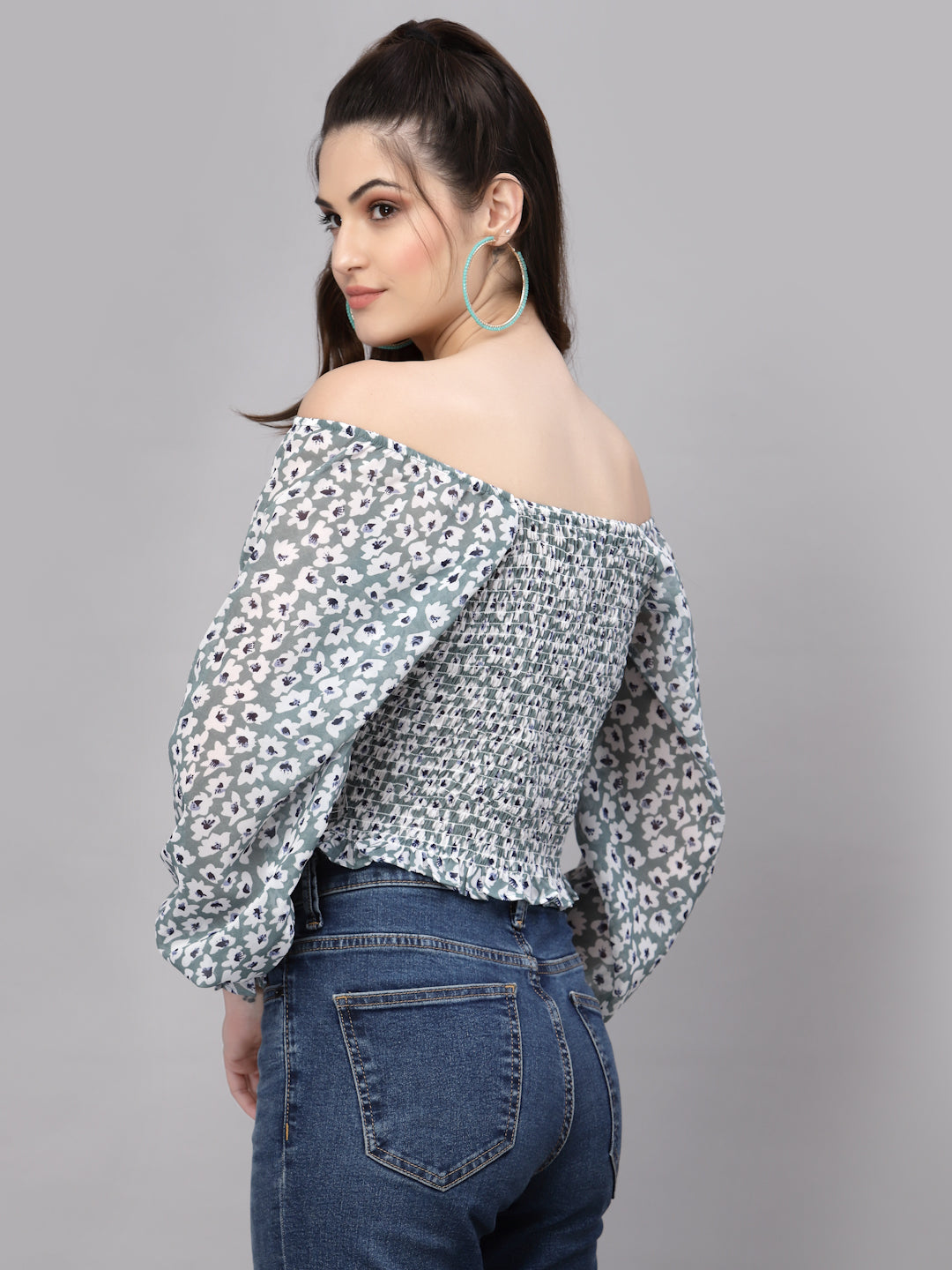 Woman wearing a green floral off-shoulder top and blue jeans on a gray background