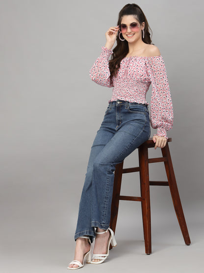 Woman wearing a pink floral top and blue jeans sitting on a wooden stool against a gray background