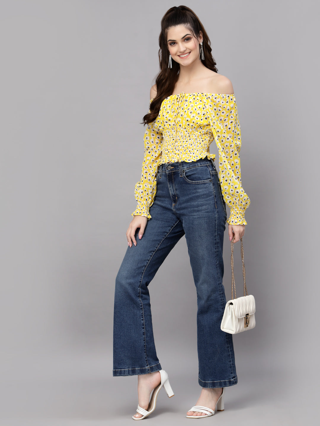 Woman wearing a yellow floral top and blue jeans, holding a white handbag on a gray background