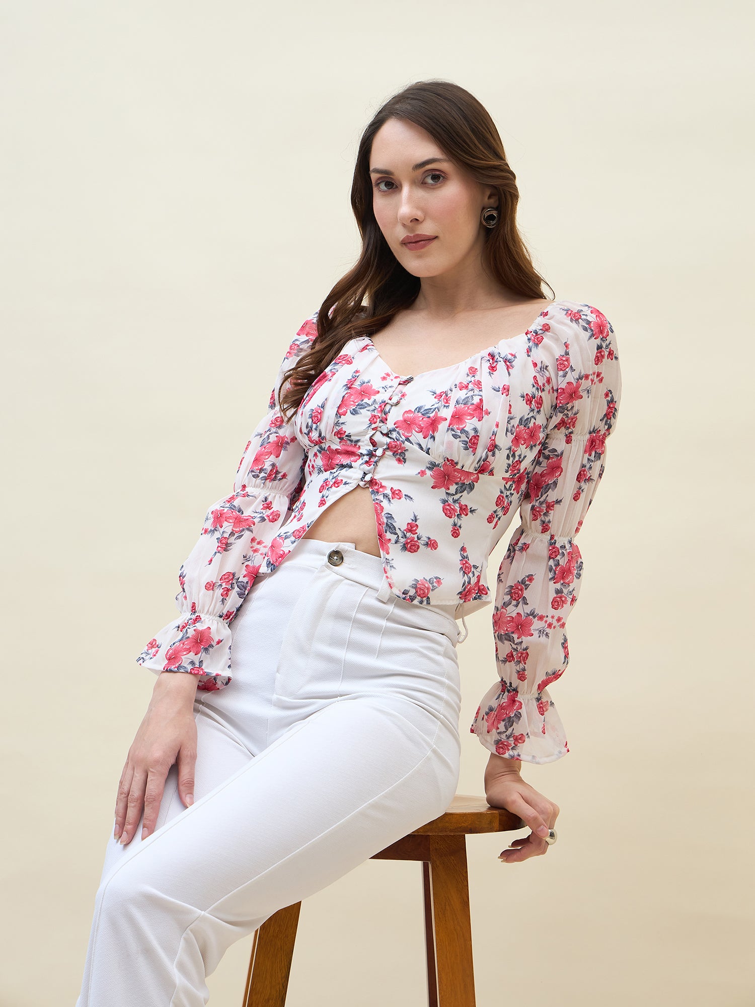 Woman wearing a white & pink floral top and white pants sitting on a stool against a beige background