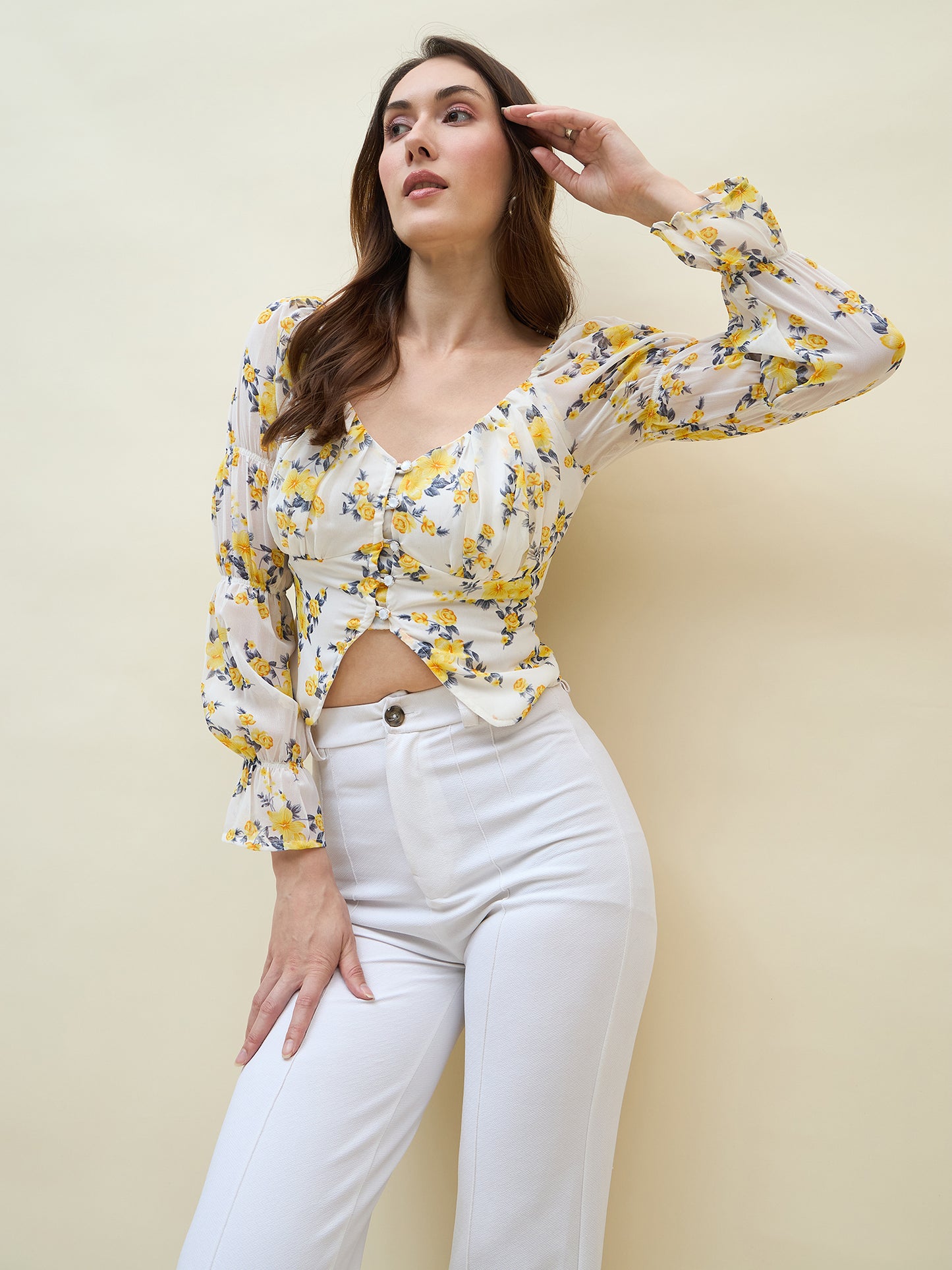 Woman wearing a white & yellow floral top and white pants against a beige background