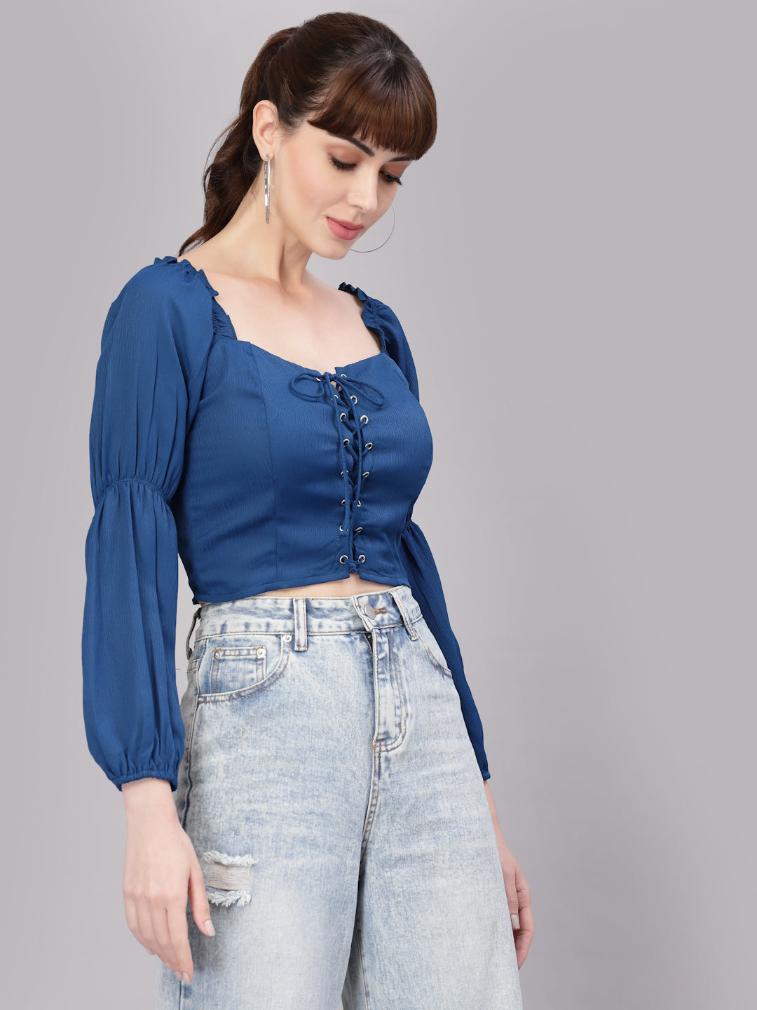 Woman wearing a blue top with lace-up front and light blue jeans on a gray background