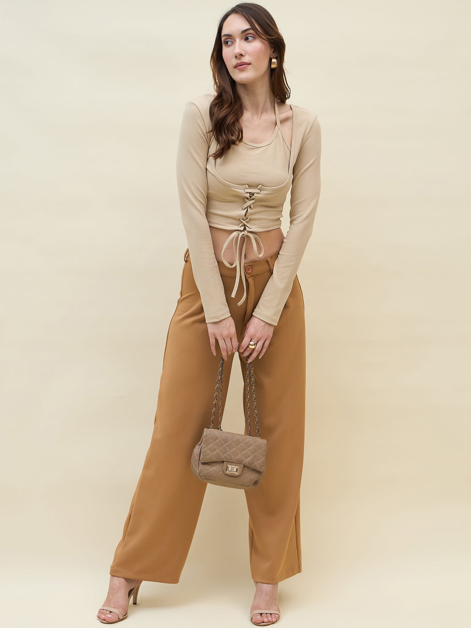Woman wearing a cream top and brown pants holding a matching handbag against a beige background