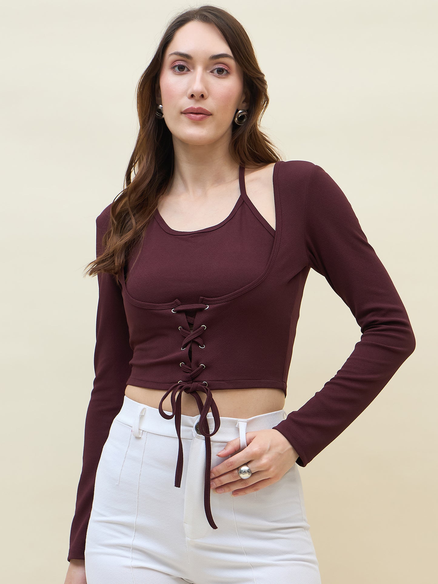 Woman wearing a wine long-sleeve crop top with lace-up front and white pants on a beige background