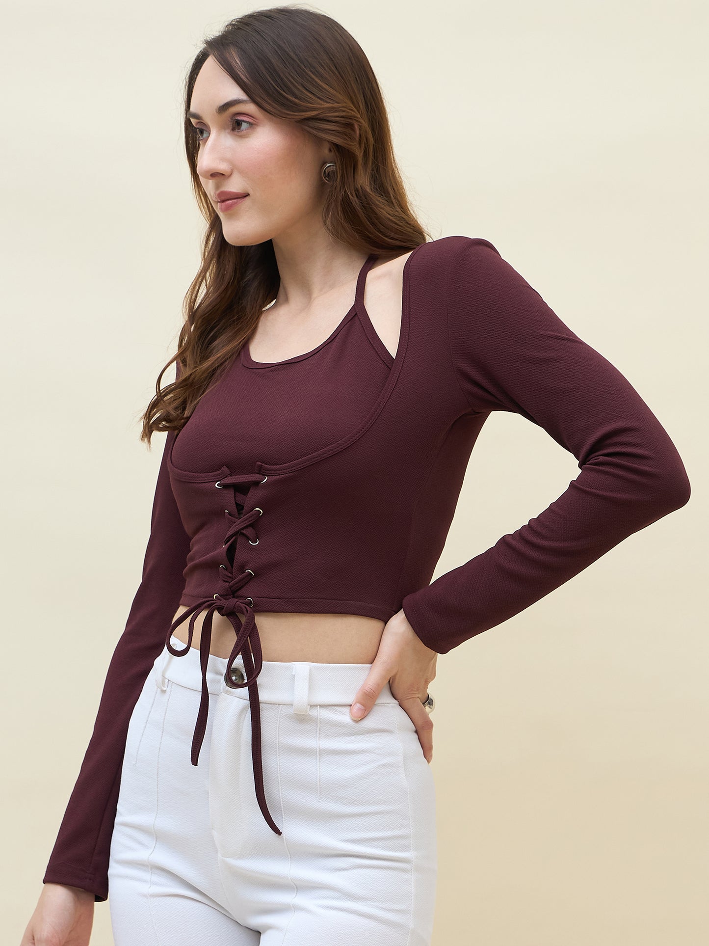 Woman wearing a maroon long-sleeve crop top with lace-up details and white pants on a beige background