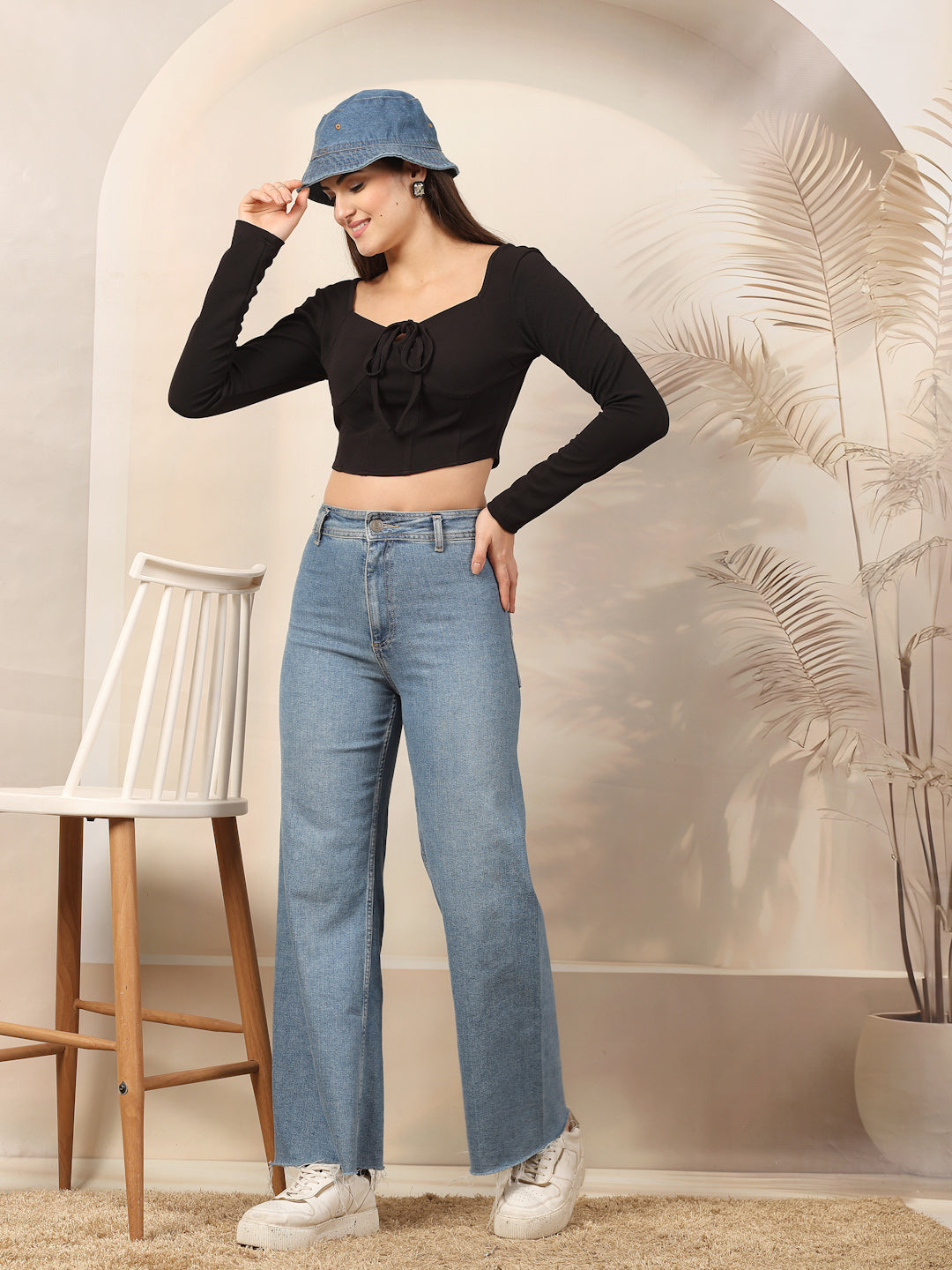 Woman wearing a black long-sleeve top and blue jeans standing in a room with a decorative wall and chair.