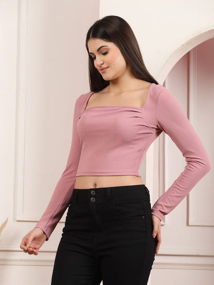 Woman wearing a mauve long-sleeve top and black pants against a light-colored wall.