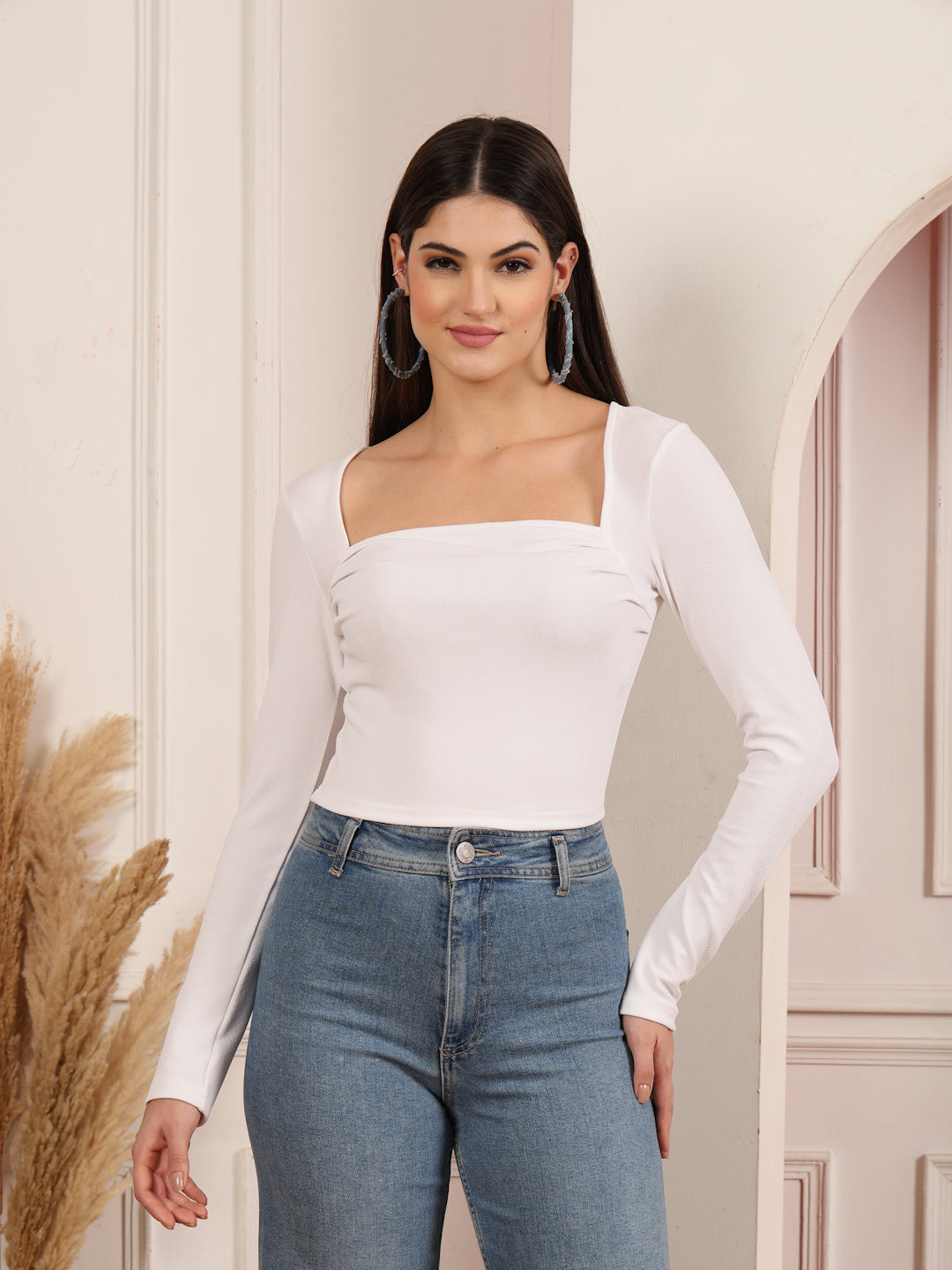 Woman wearing a white long-sleeve top and blue jeans standing against a neutral wall.