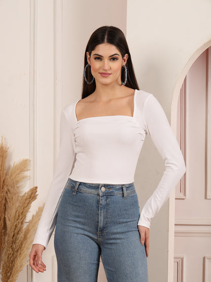 Woman wearing a white long-sleeve top and blue jeans standing against a neutral wall.