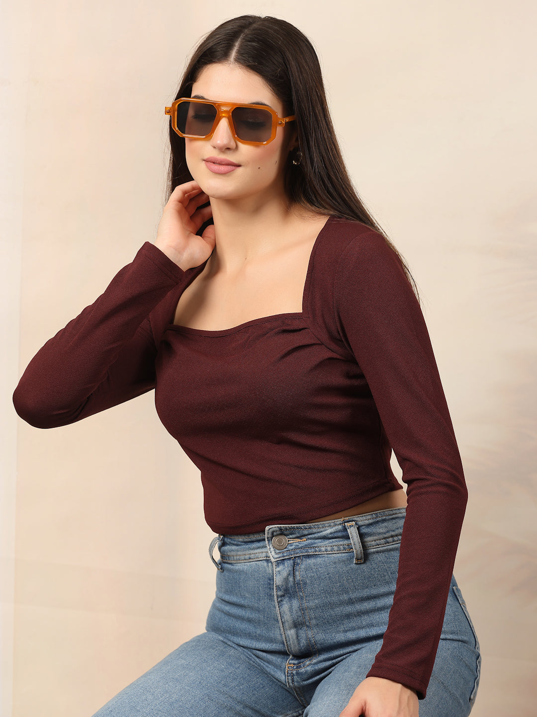 Woman wearing a wine long-sleeve top and blue jeans with sunglasses on a beige background