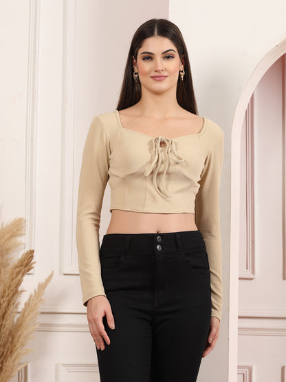 Woman wearing a beige long-sleeve top with a ruched design and black pants against a white wall.