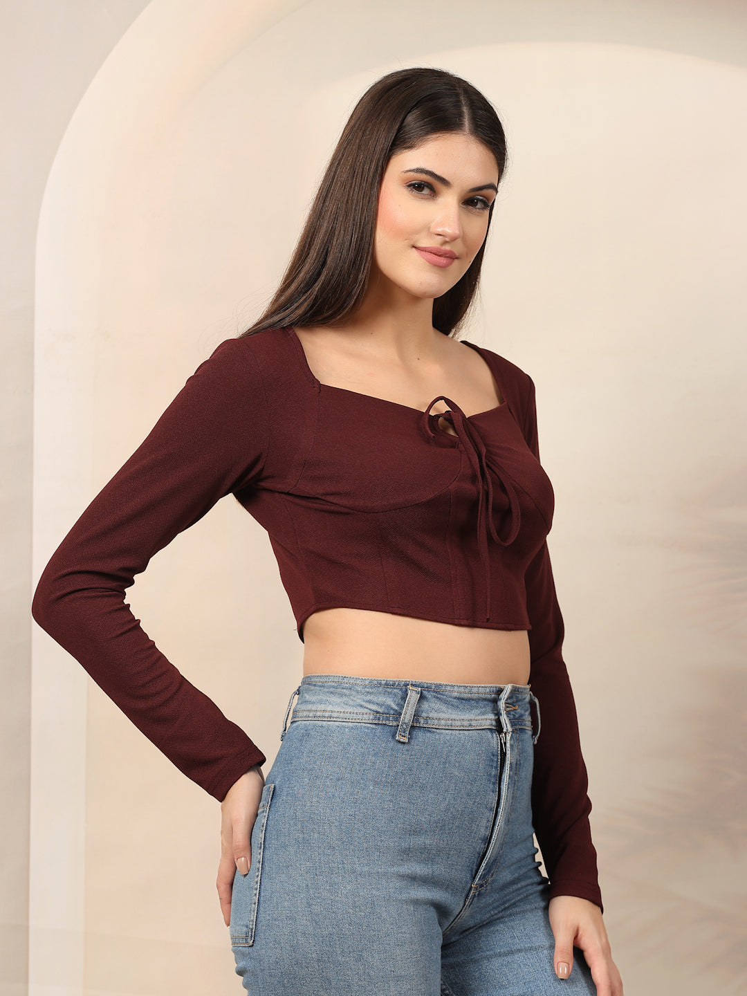Woman wearing a wine long-sleeve crop top with a tie detail and blue jeans against a beige background