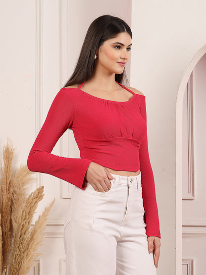 Woman wearing a pink long-sleeve top and white pants against a neutral wall.