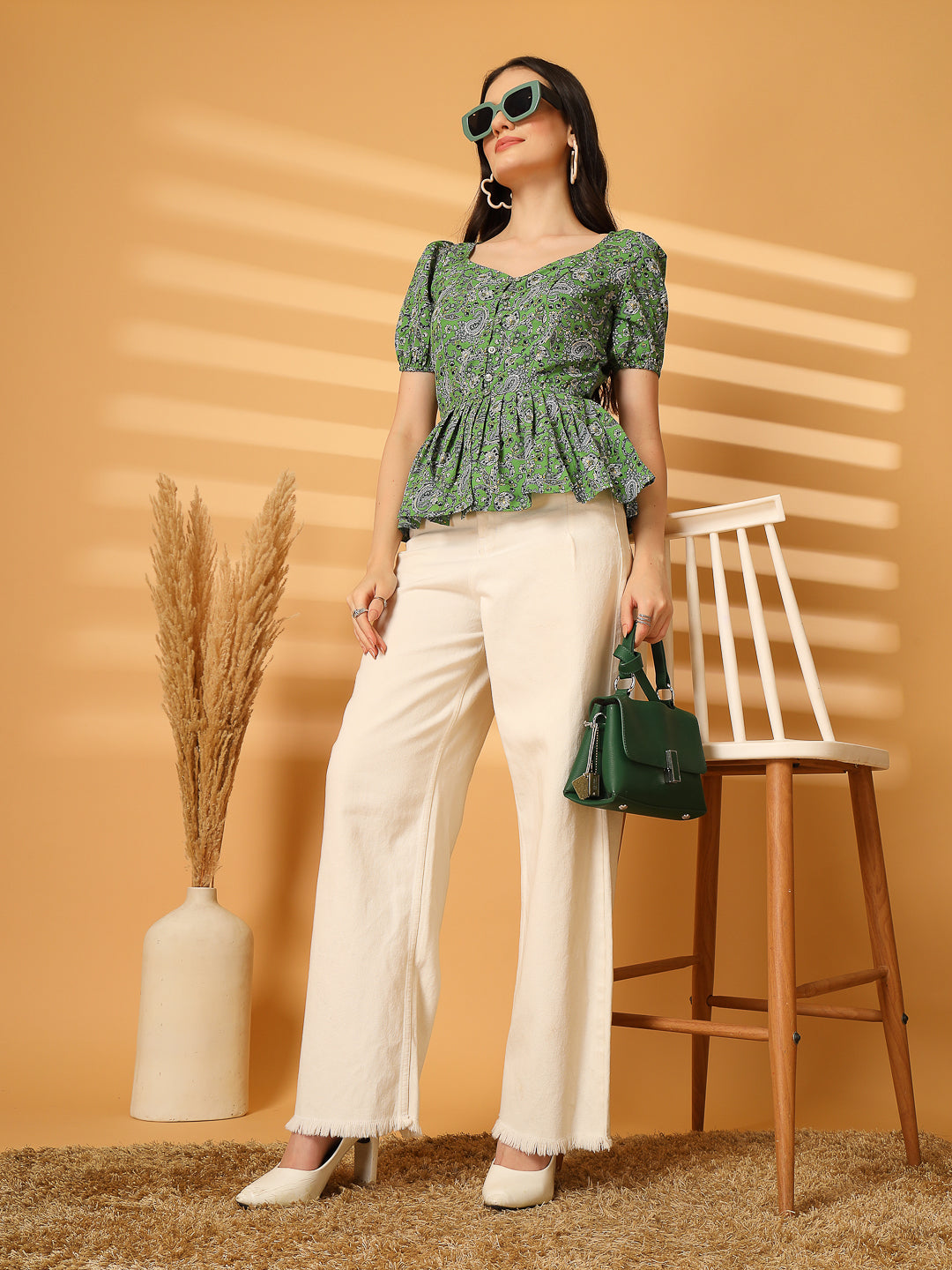 Woman in a green floral top and white pants standing against an orange wall with a chair and vase.