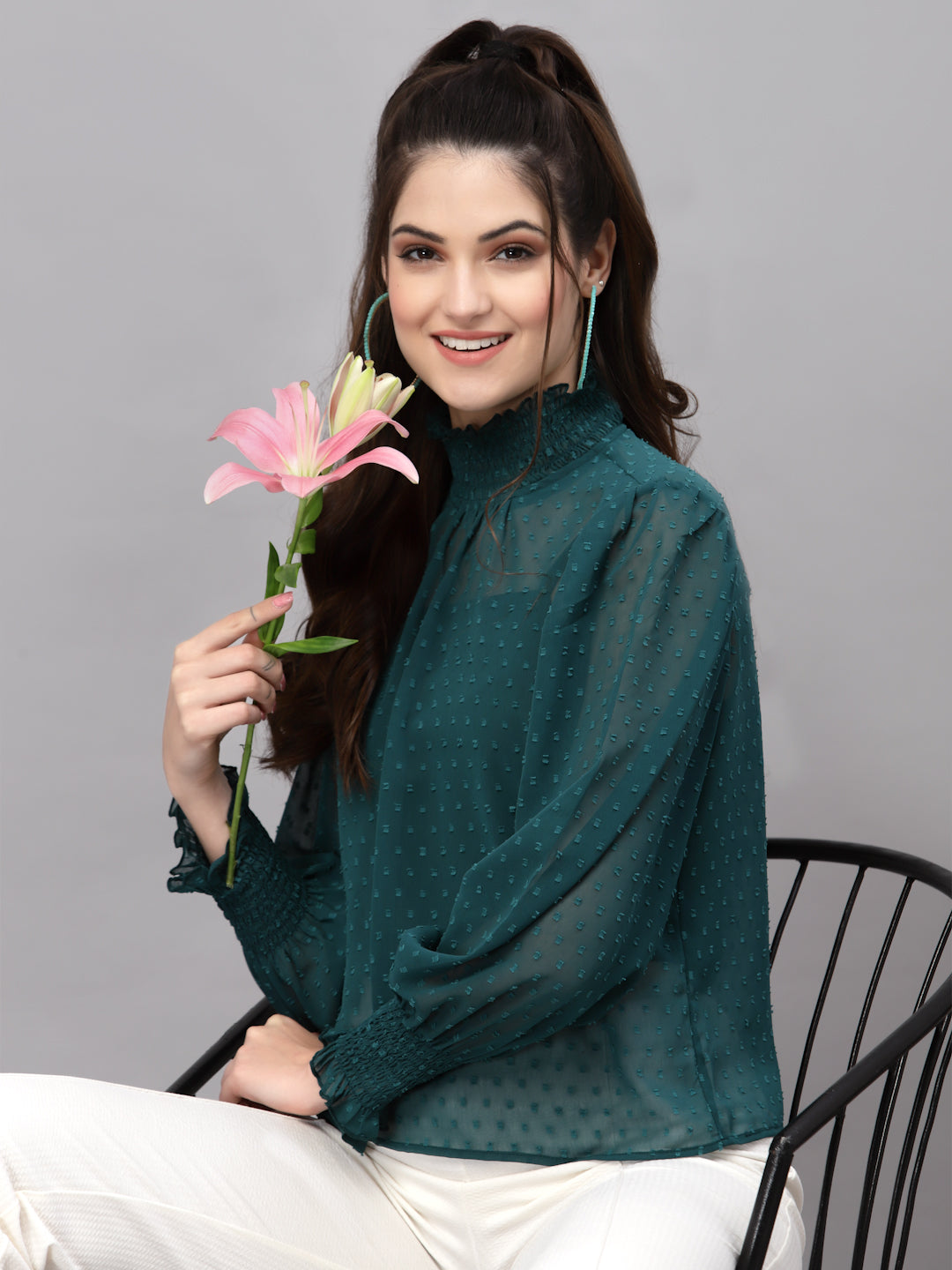 Woman holding a pink flower, wearing a green top and white pants, sitting on a chair against a gray background.