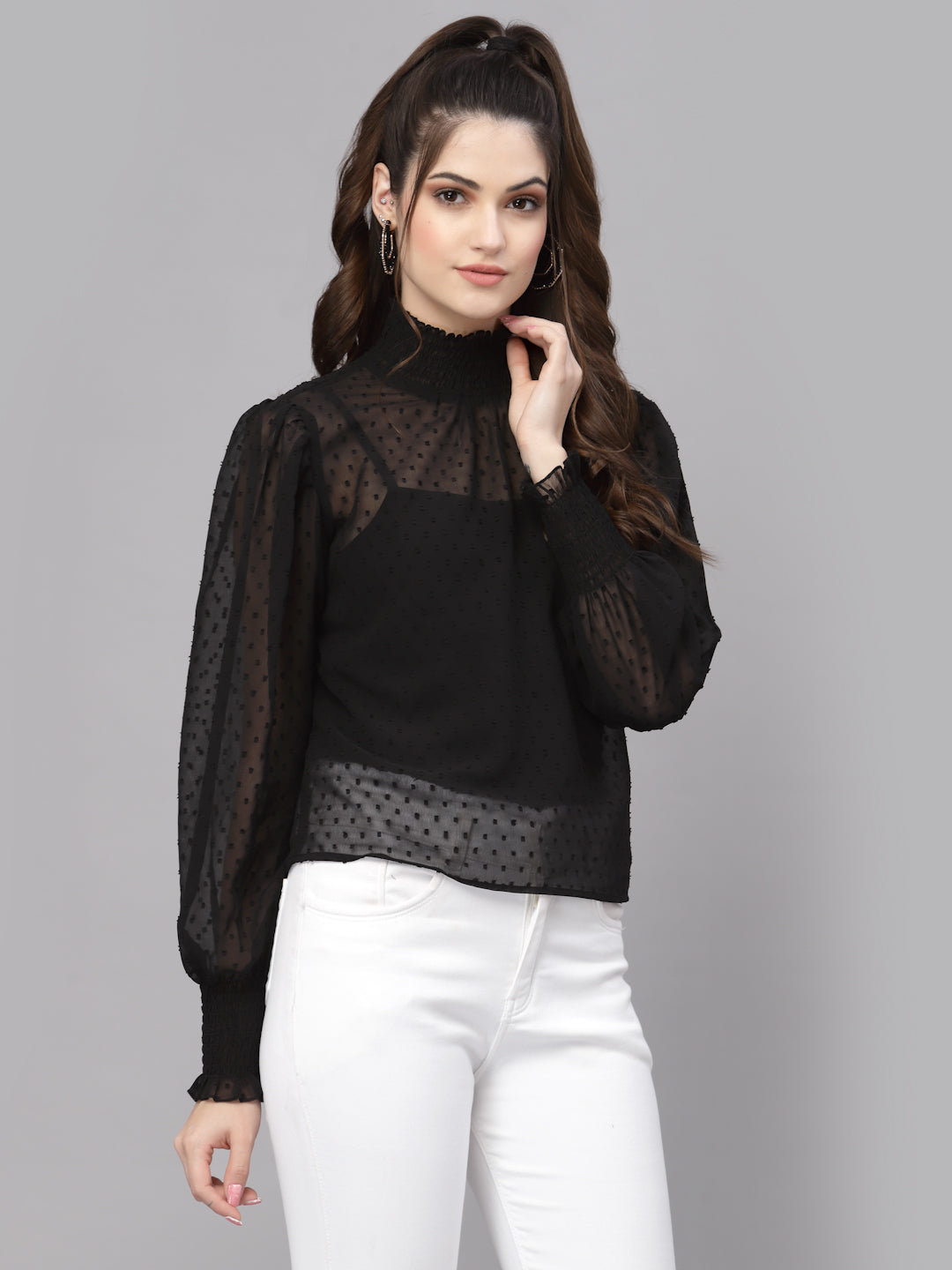 Woman wearing a black blouse with sheer sleeves and white pants on a gray background