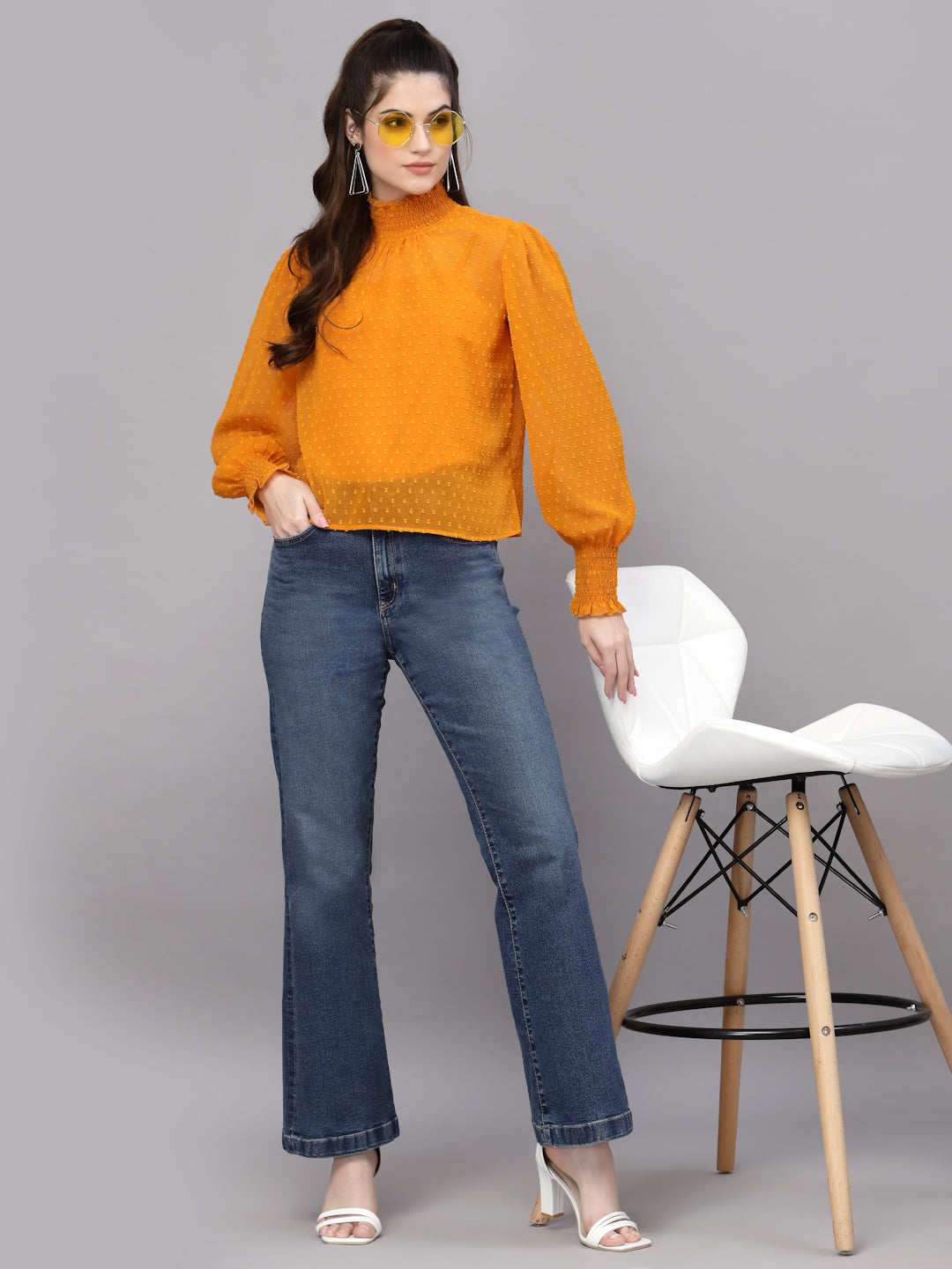 Woman wearing a mustard top and blue jeans standing next to a white chair on a gray background