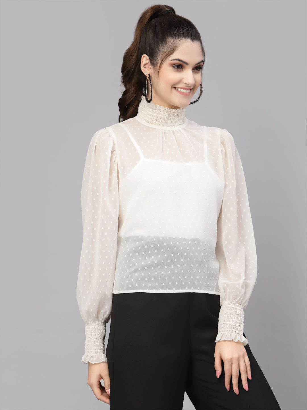 Woman wearing a sheer white top over a white top with black pants on a gray background