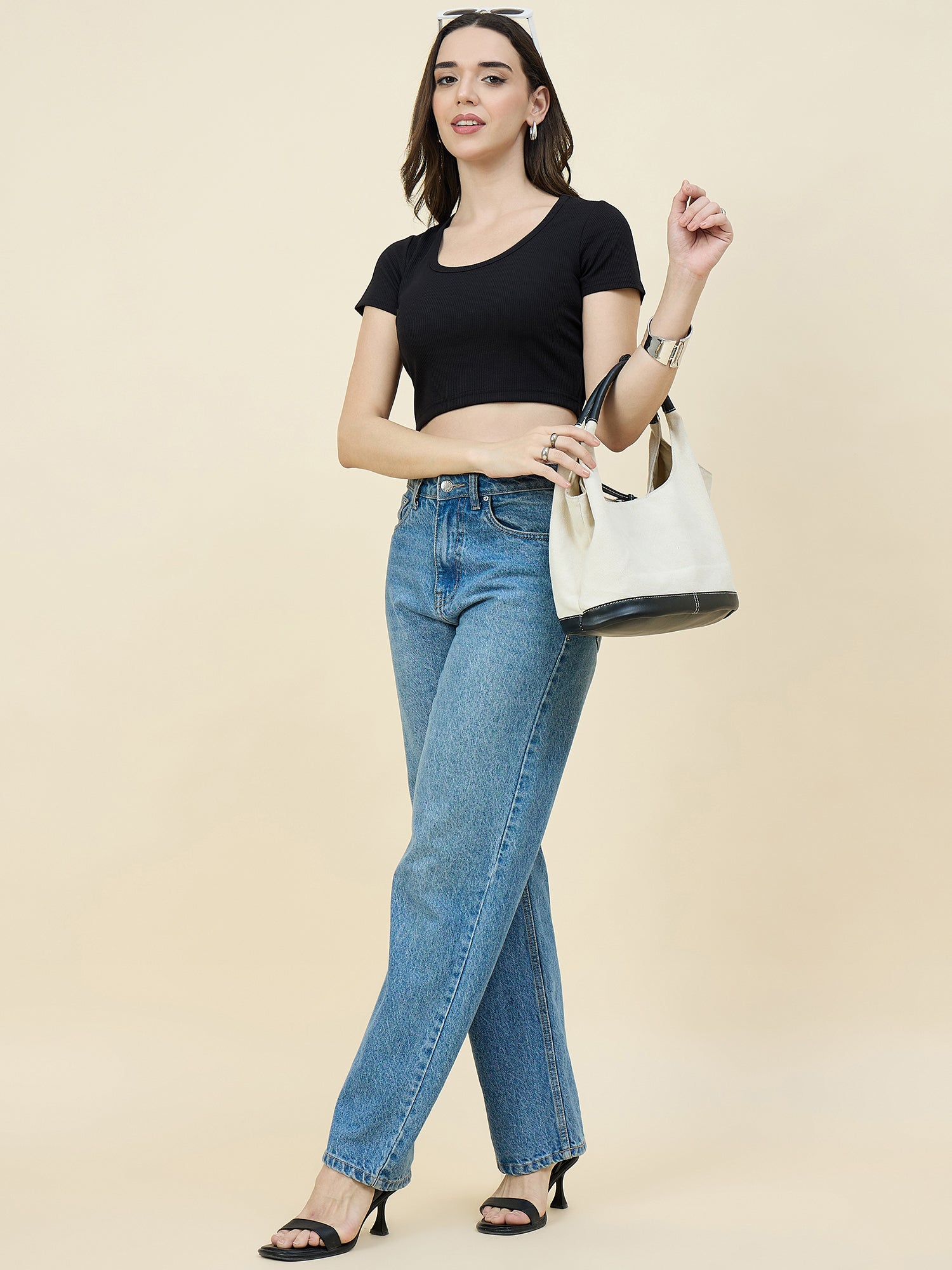 Woman wearing a black crop top and blue jeans, holding a white handbag on a beige background