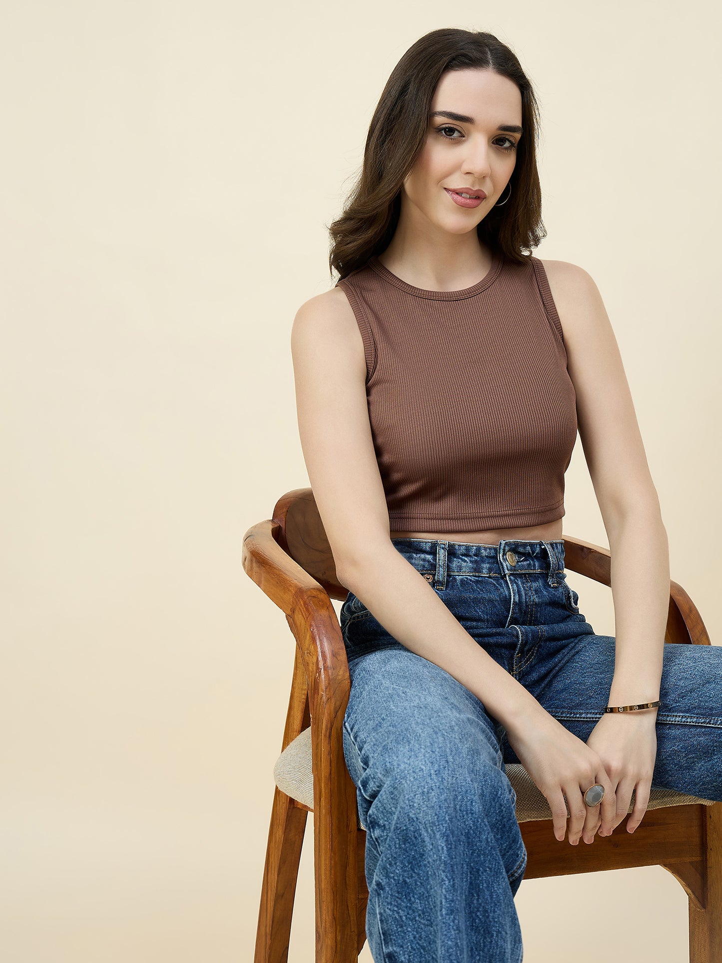 Woman wearing a brown sleeveless top and blue jeans sitting on a wooden chair against a beige background
