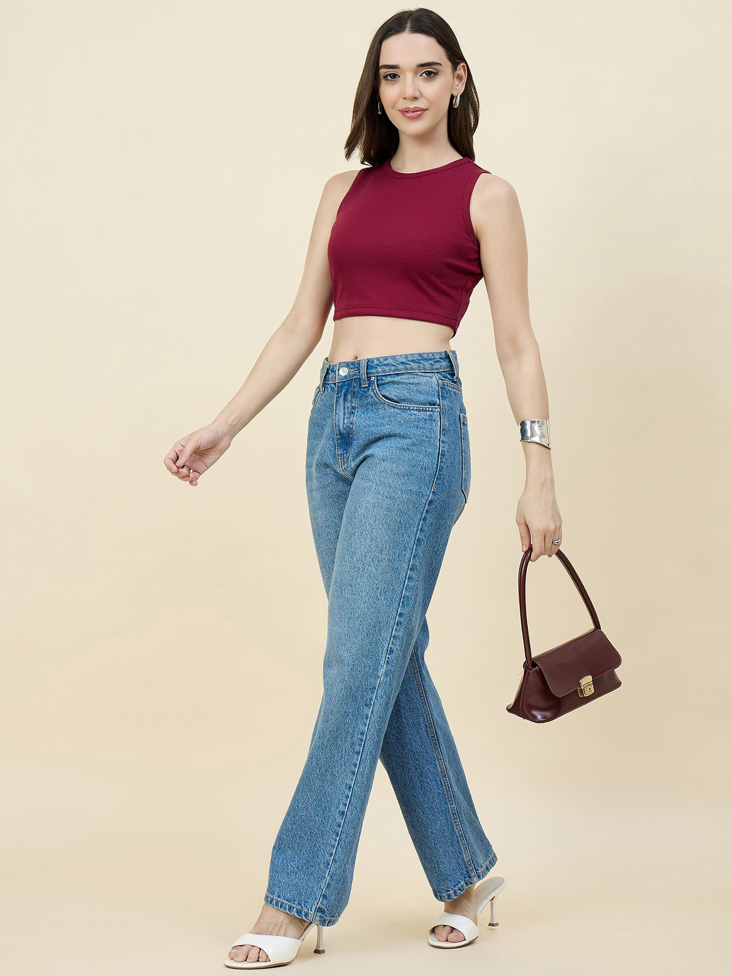 Woman wearing a maroon sleeveless top and blue jeans, holding a brown handbag on a beige background