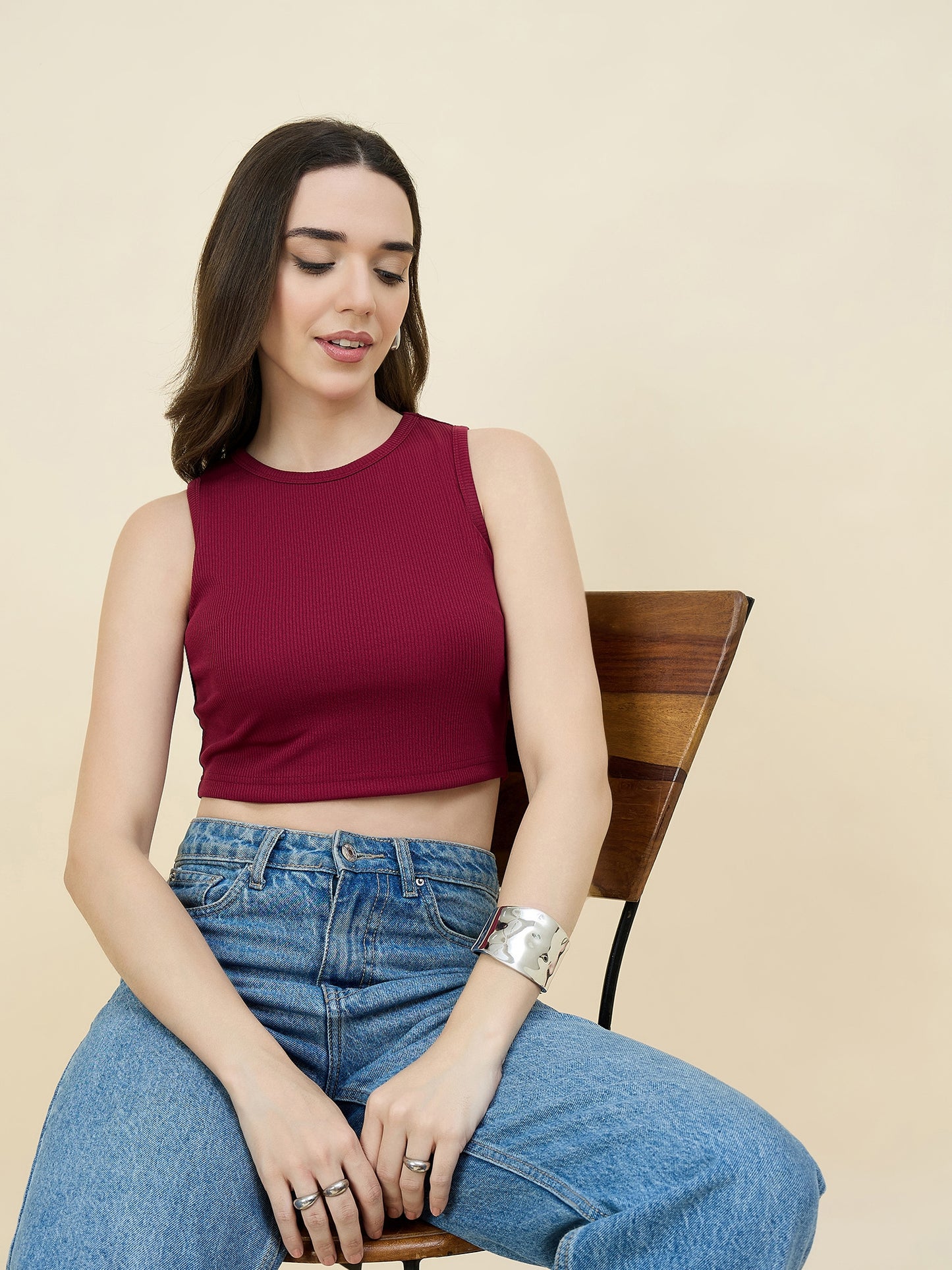 Woman wearing a maroon sleeveless top and blue jeans sitting on a wooden chair against a beige background