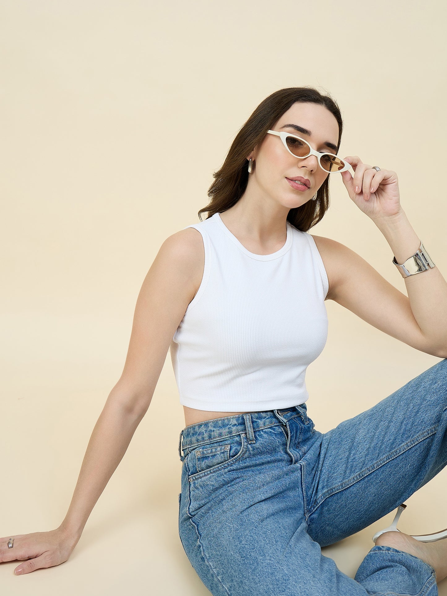 Woman wearing a white sleeveless top and blue jeans, sitting on a beige background