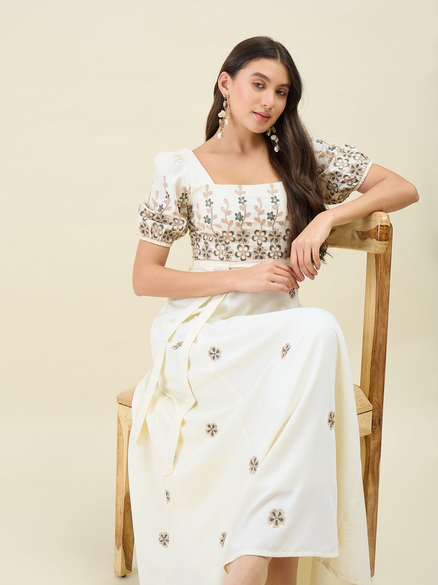 Woman in a white embroidered outfit sitting on a wooden chair against a beige background